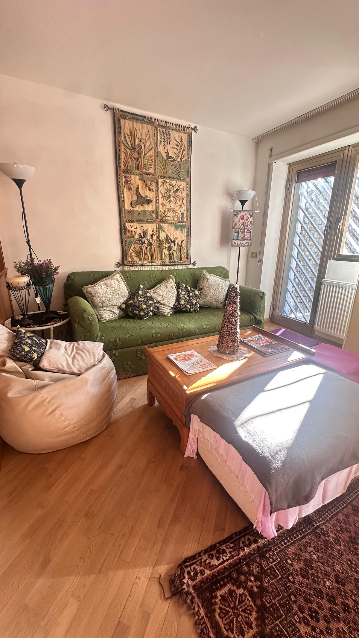 A cozy living area features a green sofa adorned with patterned cushions, complemented by a wooden coffee table stacked with magazines. A decorative wall hanging adds character, while a soft bean bag provides additional seating. Natural light streams through the glass door leading to an outdoor space.