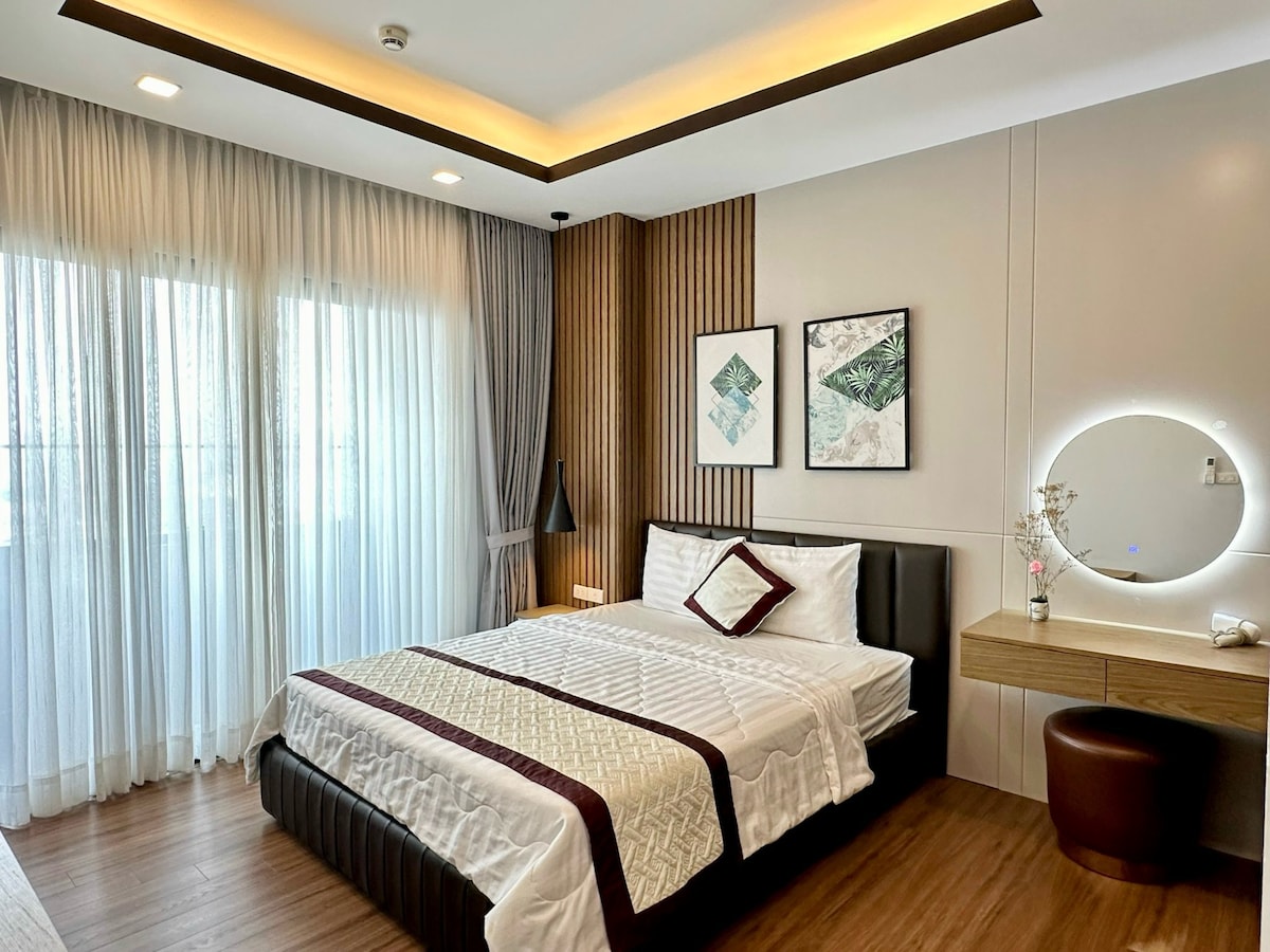 A modern bedroom features a spacious bed with a neutral-toned duvet and a decorative throw pillow. Natural light filters through sheer curtains, illuminating the space. A minimalist desk and a round mirror are positioned against a textured wall, complemented by framed artwork.