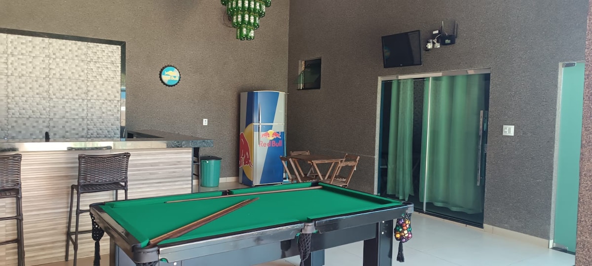 A spacious entertainment area features a green felt pool table with cue sticks resting on its edge. A modern bar with stools is positioned against the back wall, adorned with decorative lighting. Large glass doors lead to the outside, enhancing the openness of the space.