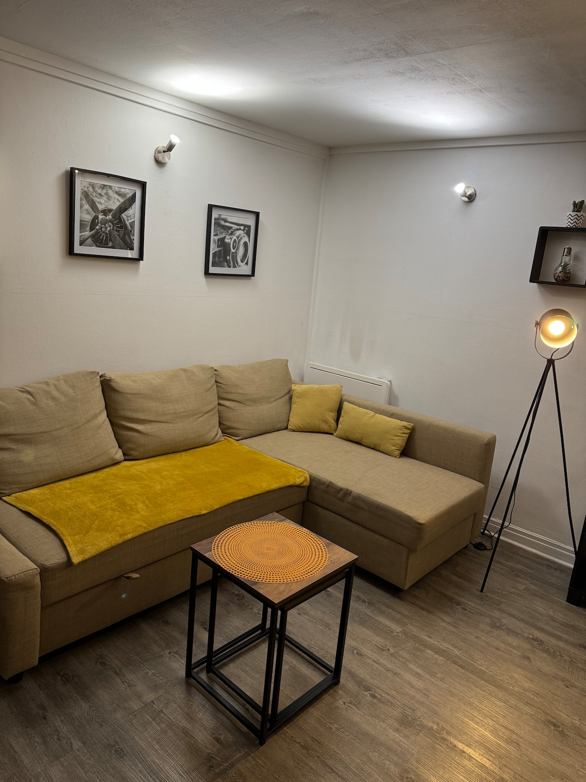 A cozy seating area is presented with a beige sectional sofa, complemented by a yellow throw. A small wooden table with a textured surface is placed in front, and framed artwork is displayed on the wall. Soft lighting from wall fixtures enhances the warm ambiance.