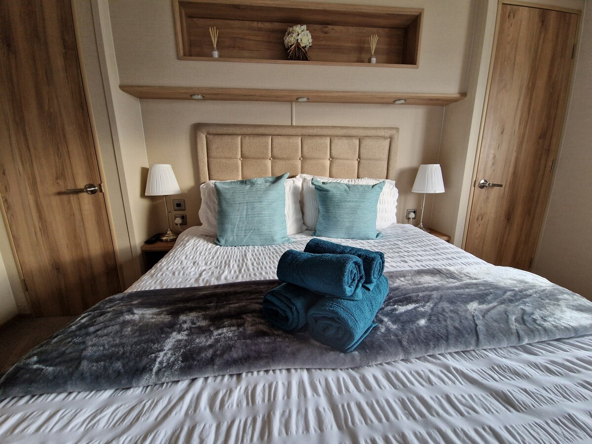 A comfortable sleeping area features a soft upholstered headboard and two decorative pillows. Freshly rolled towels are arranged neatly on the bed, complemented by a cozy throw blanket. Warm wood accents and bedside lamps create a welcoming environment.