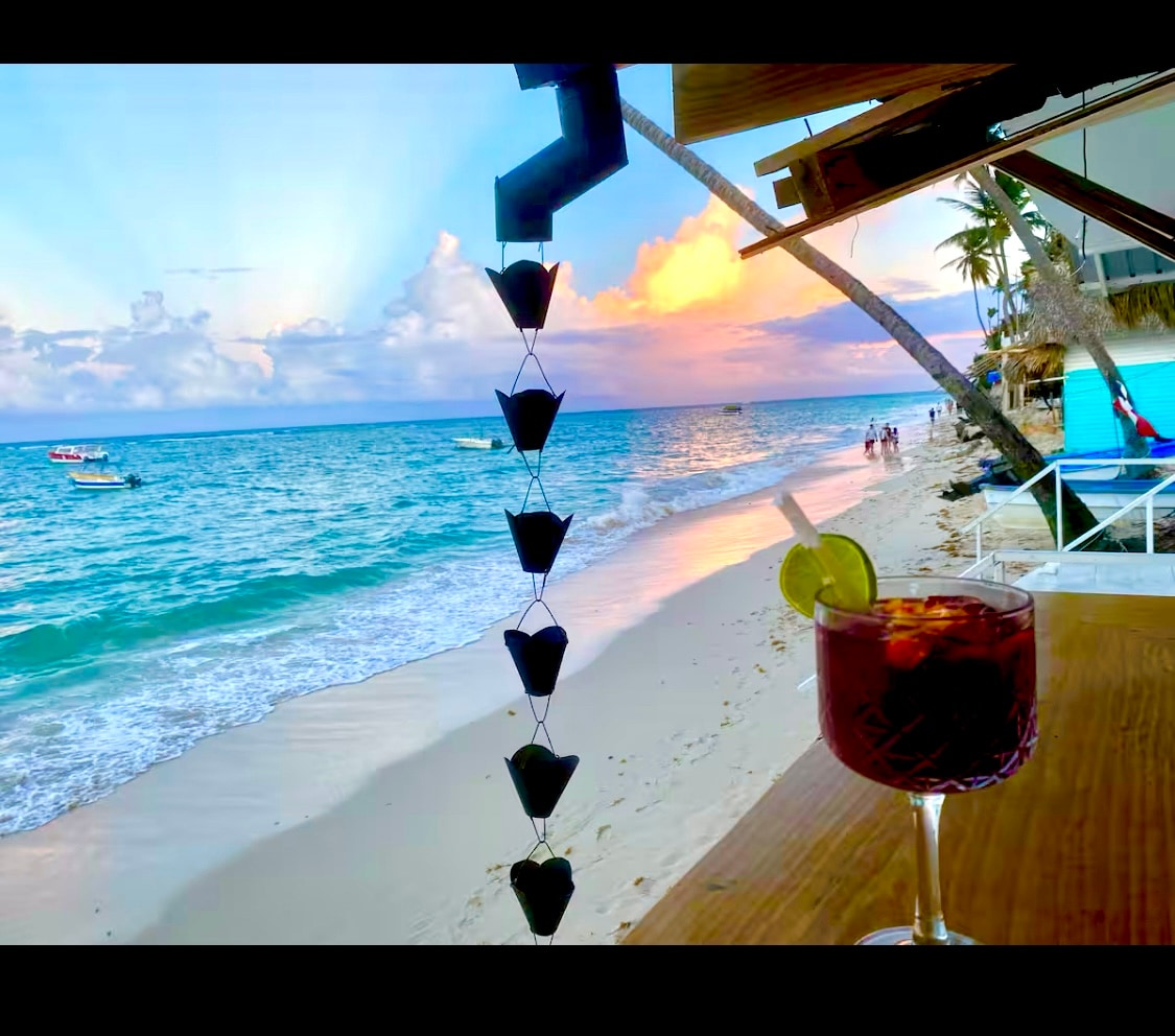 A coastal scene is captured, showcasing gentle waves lapping against a sandy shore. A beverage sits on a wooden surface, framed by a decorative element. The sky is filled with soft hues as the sun sets, while beachgoers enjoy the serene atmosphere.
