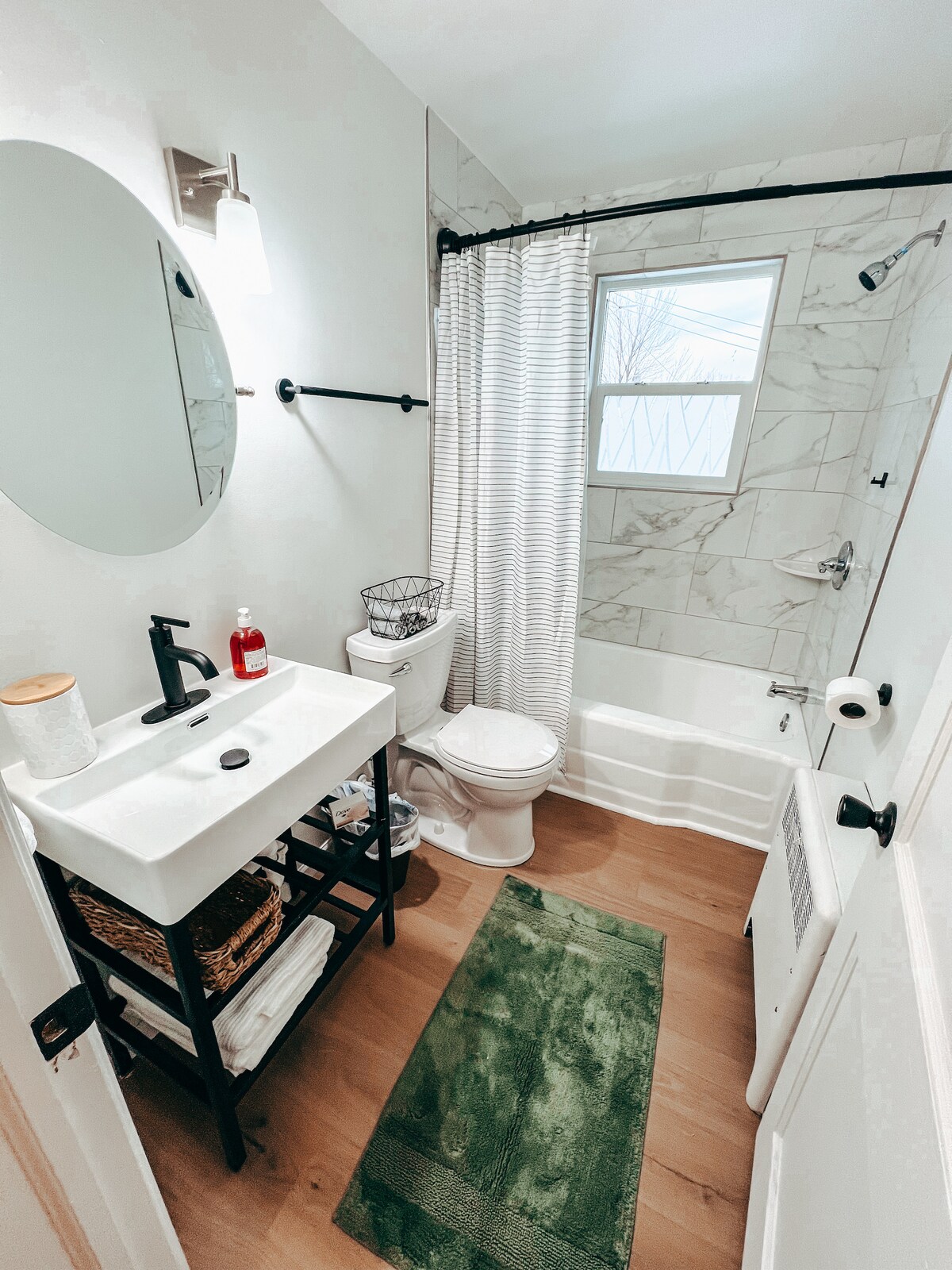 A newly renovated bathroom features a sleek tile shower and a bathtub enclosed by a shower curtain. A modern sink sits on a black metal stand, accompanied by storage baskets. Neutral tones are complemented by a green bath mat on the wooden floor and a circular mirror above the sink.