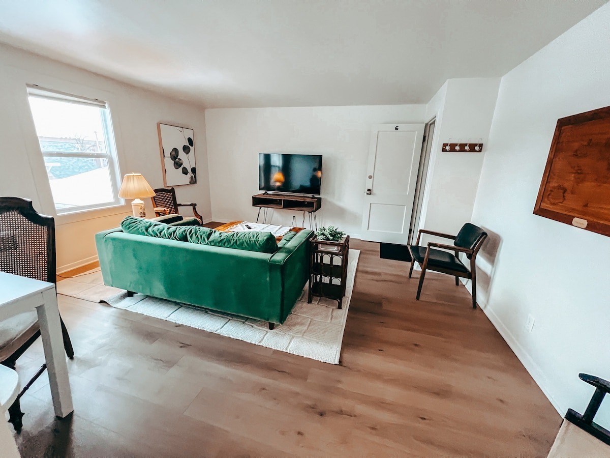 A living area features a green sofa facing a wall-mounted television. Sunlight fills the space through a large window, highlighting light wood flooring. A table lamp sits beside the sofa, and a plant adds a touch of greenery. A door leads to an entryway.