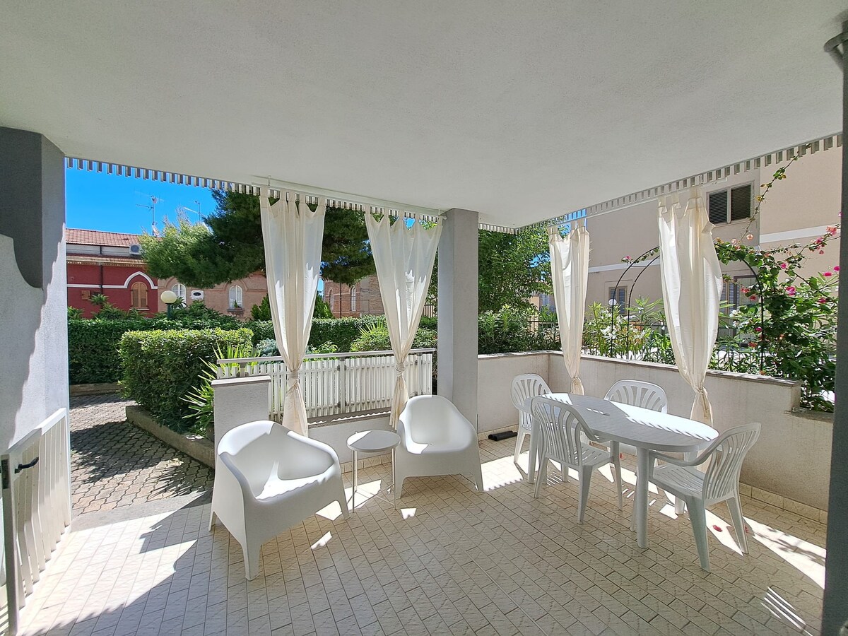An outdoor terrace features a dining table surrounded by four white chairs. Two cushioned chairs and a small table are also present. Sheer curtains gently frame the area, providing shade while allowing natural light to filter through.