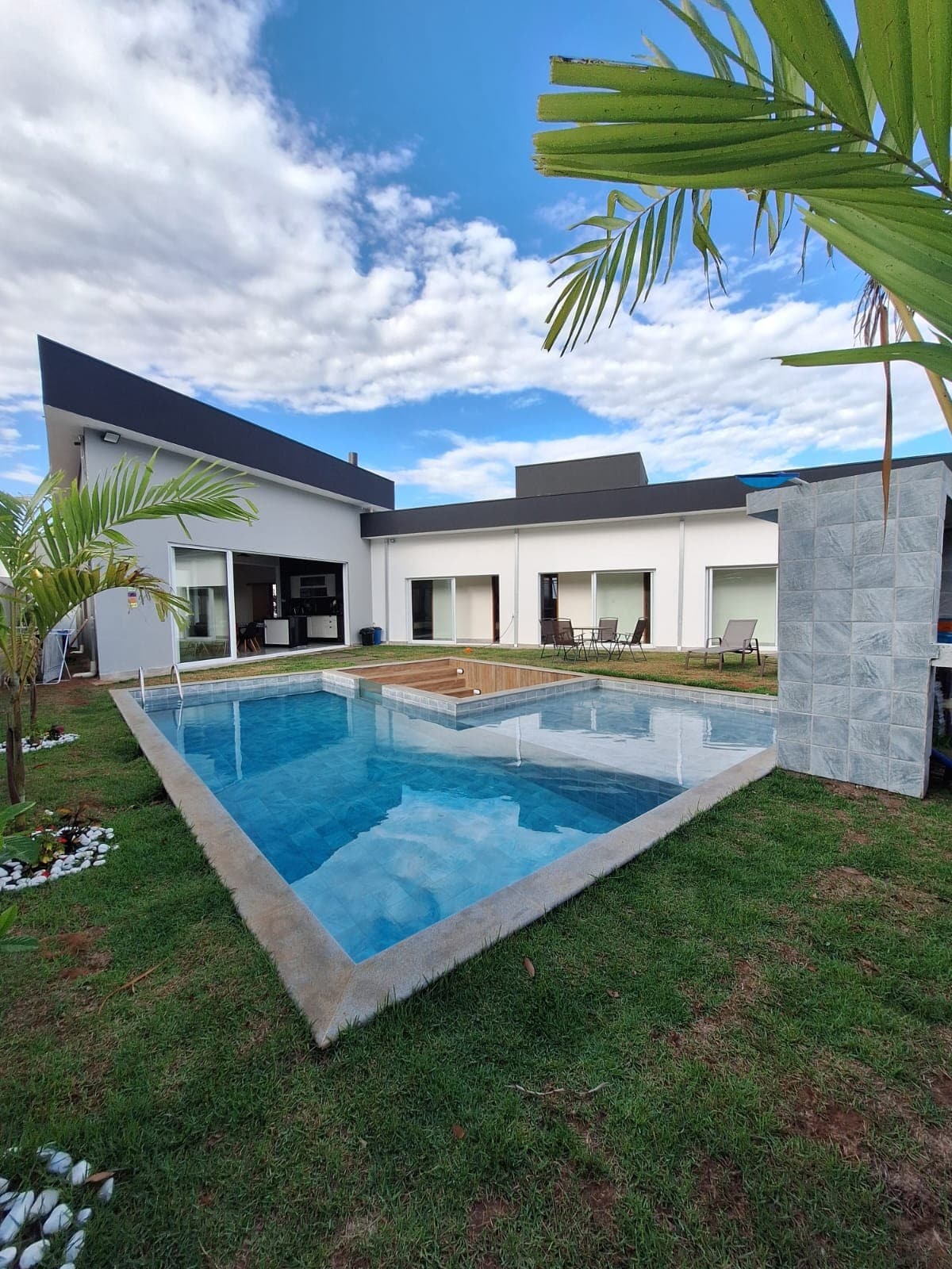 A modern house is surrounded by a lush green lawn and features a triangular swimming pool. Clear water reflects the sky, while patio furniture lines the edge. The structure showcases large windows and a contemporary roof design, ensuring ample natural light.