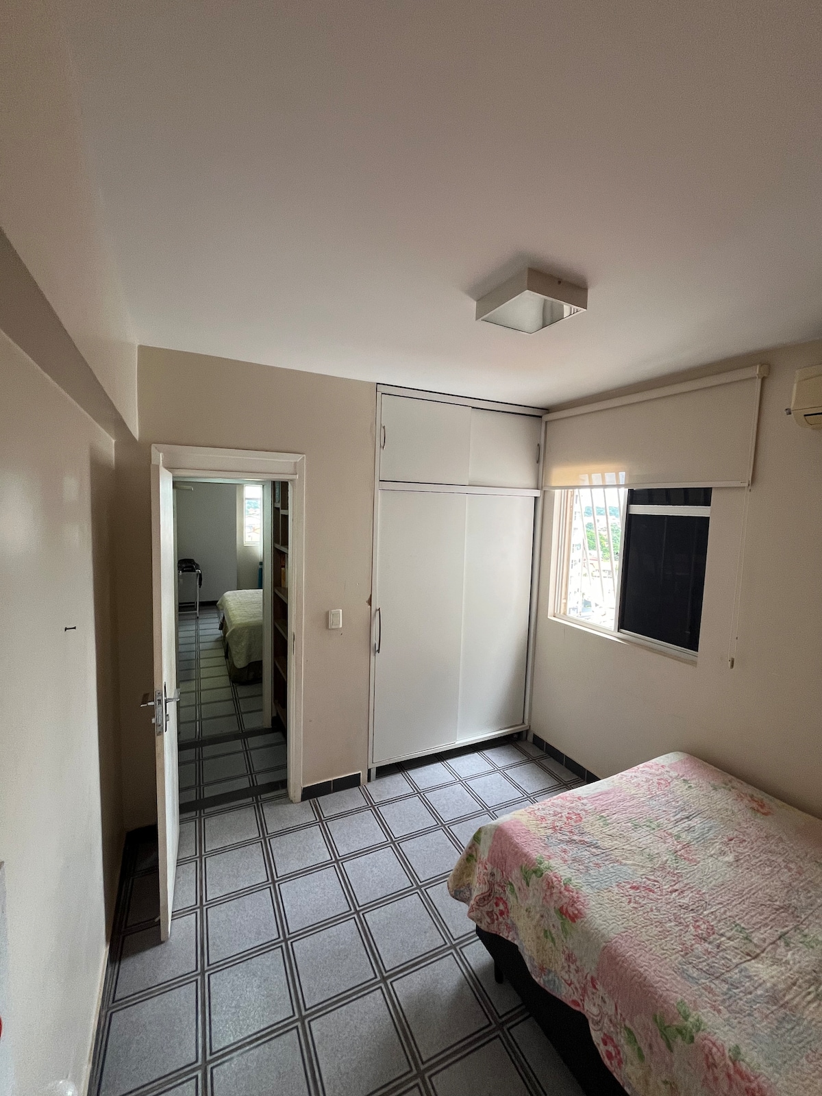 A comfortable room features a spacious bed, dressed in a floral coverlet. Natural light enters through a window, while a light fixture illuminates the ceiling. A closet with sliding doors offers storage space, and a doorway leads to an adjacent room.