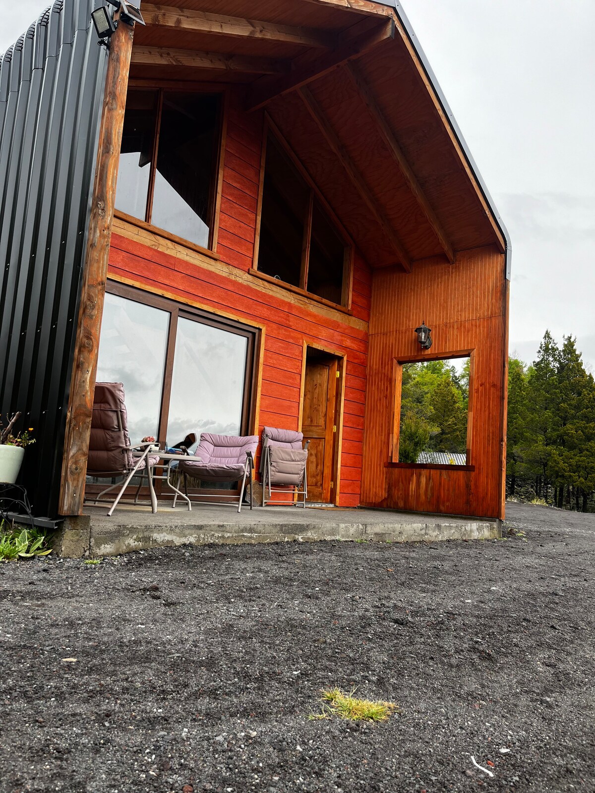A wooden cabin with a steeply pitched roof is shown, featuring large glass windows and a wooden door. A small patio area includes two comfortable chairs and a table, set on a gravel surface. Greenery is visible in the background, enhancing the natural surroundings.