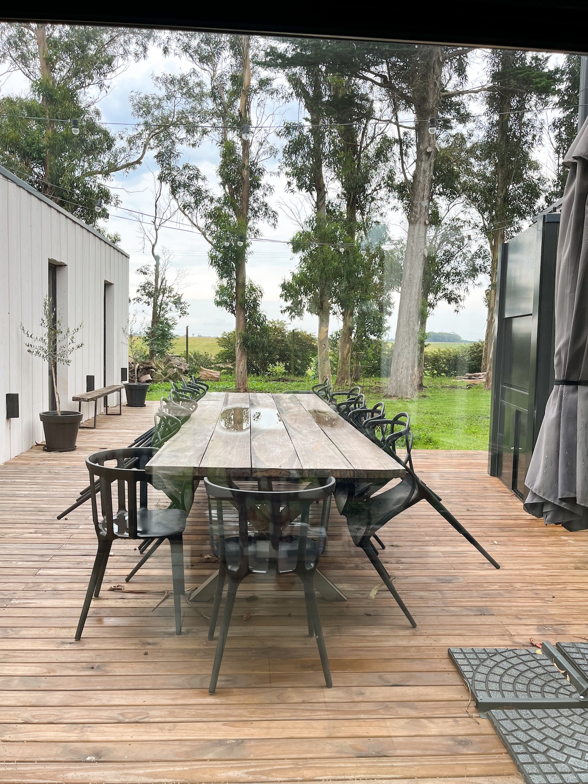 An outdoor dining area features a long wooden table surrounded by several chairs. The table is set on a wooden deck, with views of greenery in the background and tall trees lining the space. Natural light filters through the glass doors.
