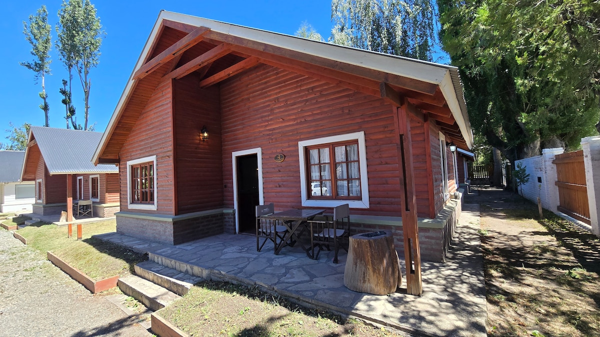A wooden cabin with a pitched roof is showcased in this image. It features multiple windows that allow natural light to enter. Outdoor seating is provided at a stone-paved entrance, alongside a wooden stump. Surrounding greenery adds to the serene environment.