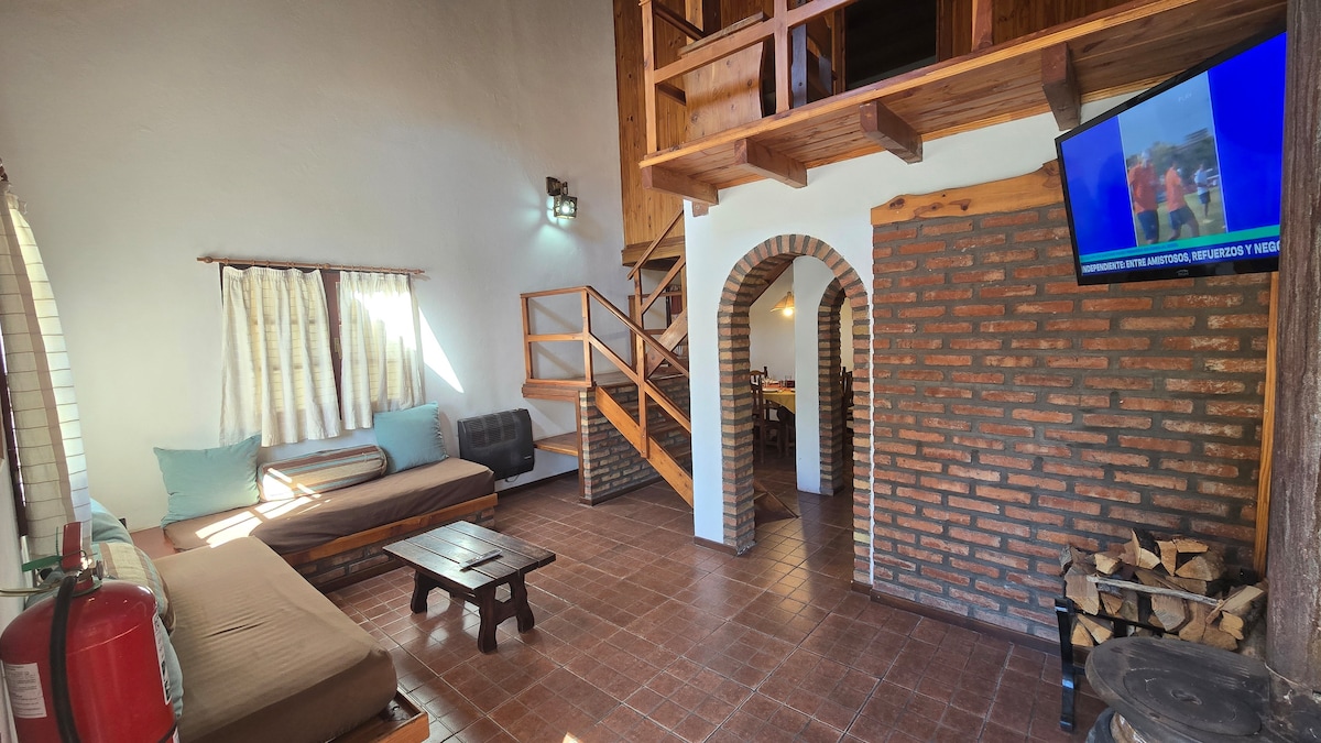 The living area showcases a mix of textures, featuring a wooden staircase leading to the upper floor. A television is mounted on a brick wall, while a cozy seating area includes a modern couch and a wooden coffee table. Sunlight enters through large windows, illuminating the space.