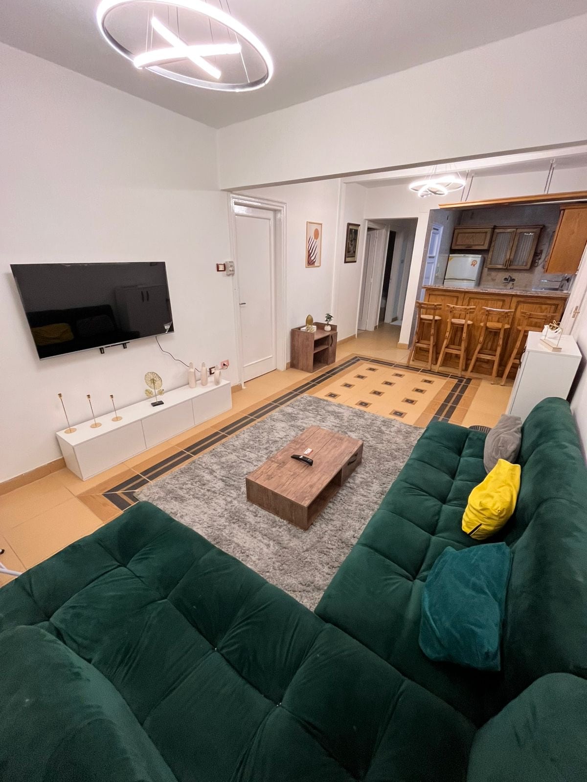 A spacious living area is highlighted by a green sectional sofa with decorative pillows. A wooden coffee table and a television on a white media console are centered on a plush gray rug. The background shows a dining area with bar stools and an open kitchen.