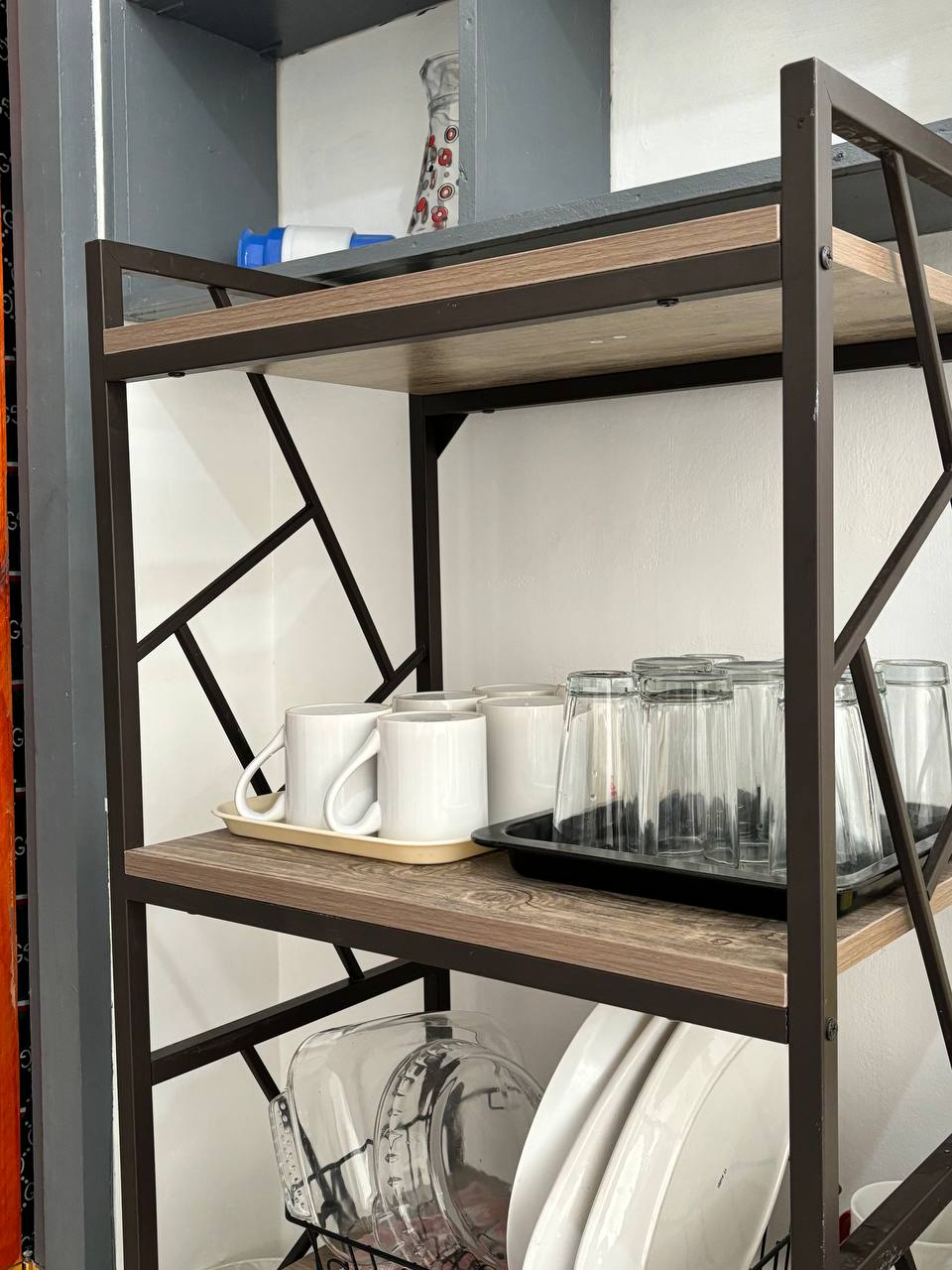 A sturdy metal and wood shelving unit displays various kitchen items, including white mugs and glass containers. A dark tray holds additional glassware, while neatly arranged dinner plates and clear bowls occupy the lower shelves, providing easy access and organization.