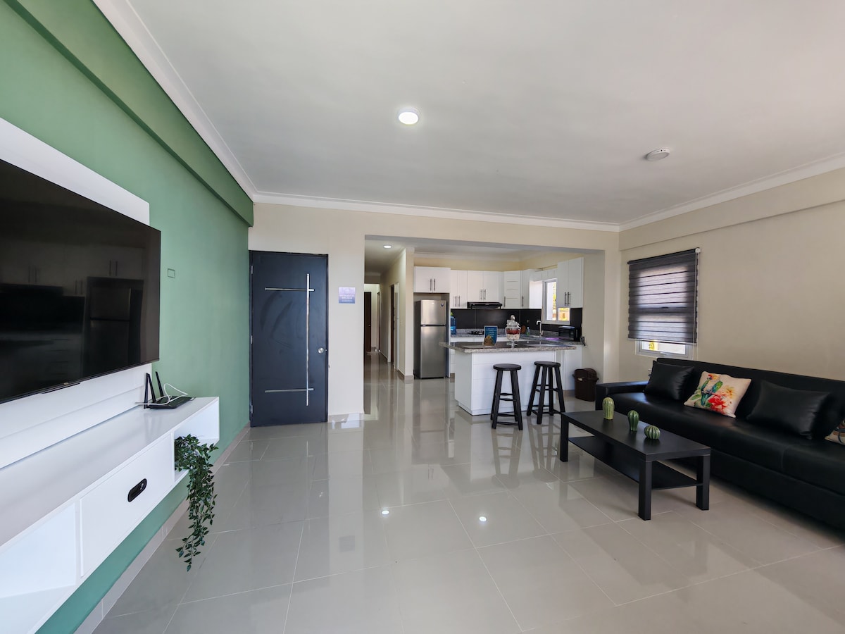 The spacious living area features a modern sectional sofa and a sleek coffee table. A flat-screen TV is mounted on the wall, with a vibrant wall art piece adding a splash of color. The kitchen is visible in the background, enhancing the open-concept layout.