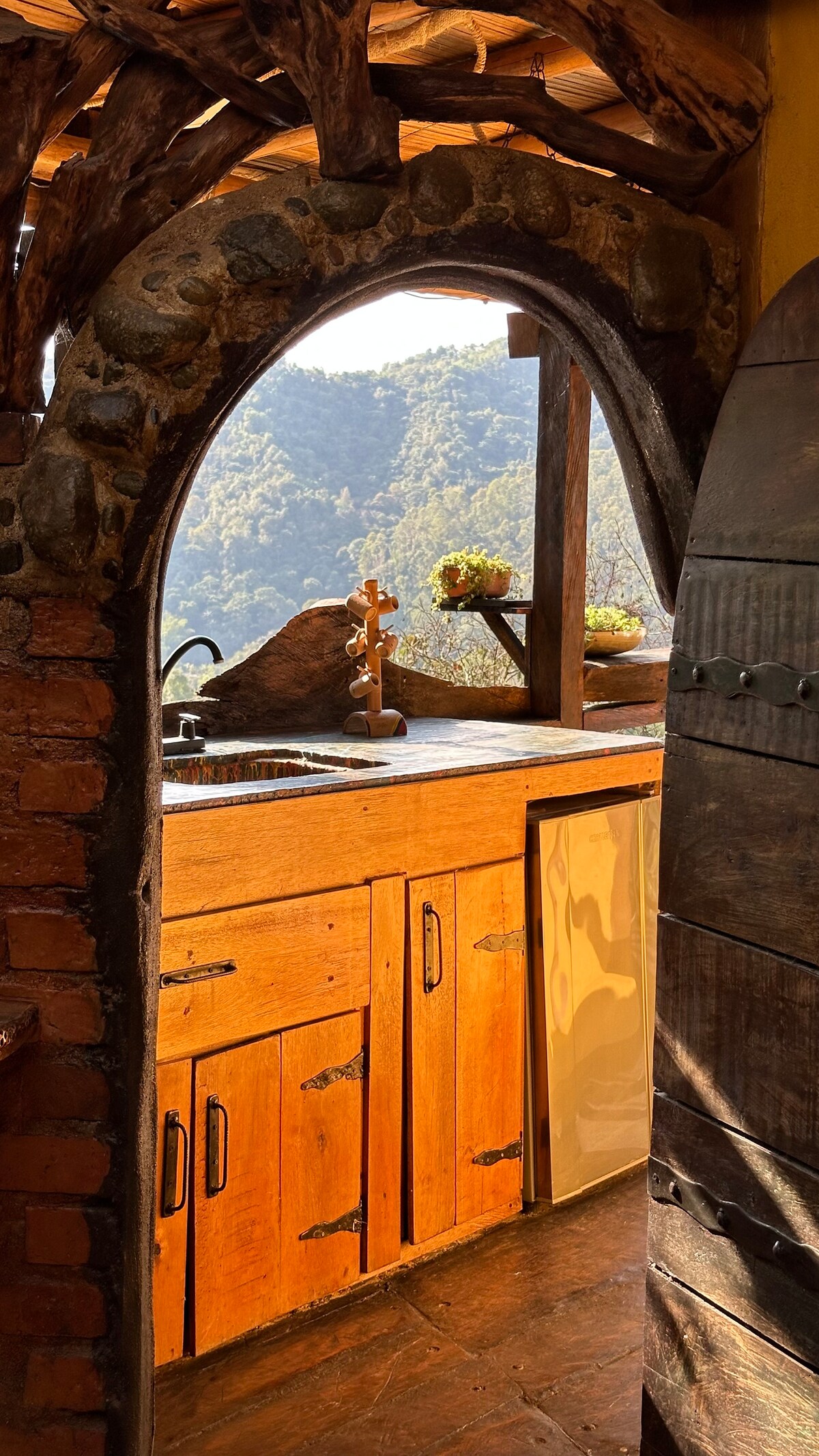 A rustic kitchen area is framed by an arched stone doorway, highlighting warm wooden cabinetry and a scenic view of the mountains through an open window. Natural light floods the space, illuminating the stone and wood contrasts, creating a cozy atmosphere.