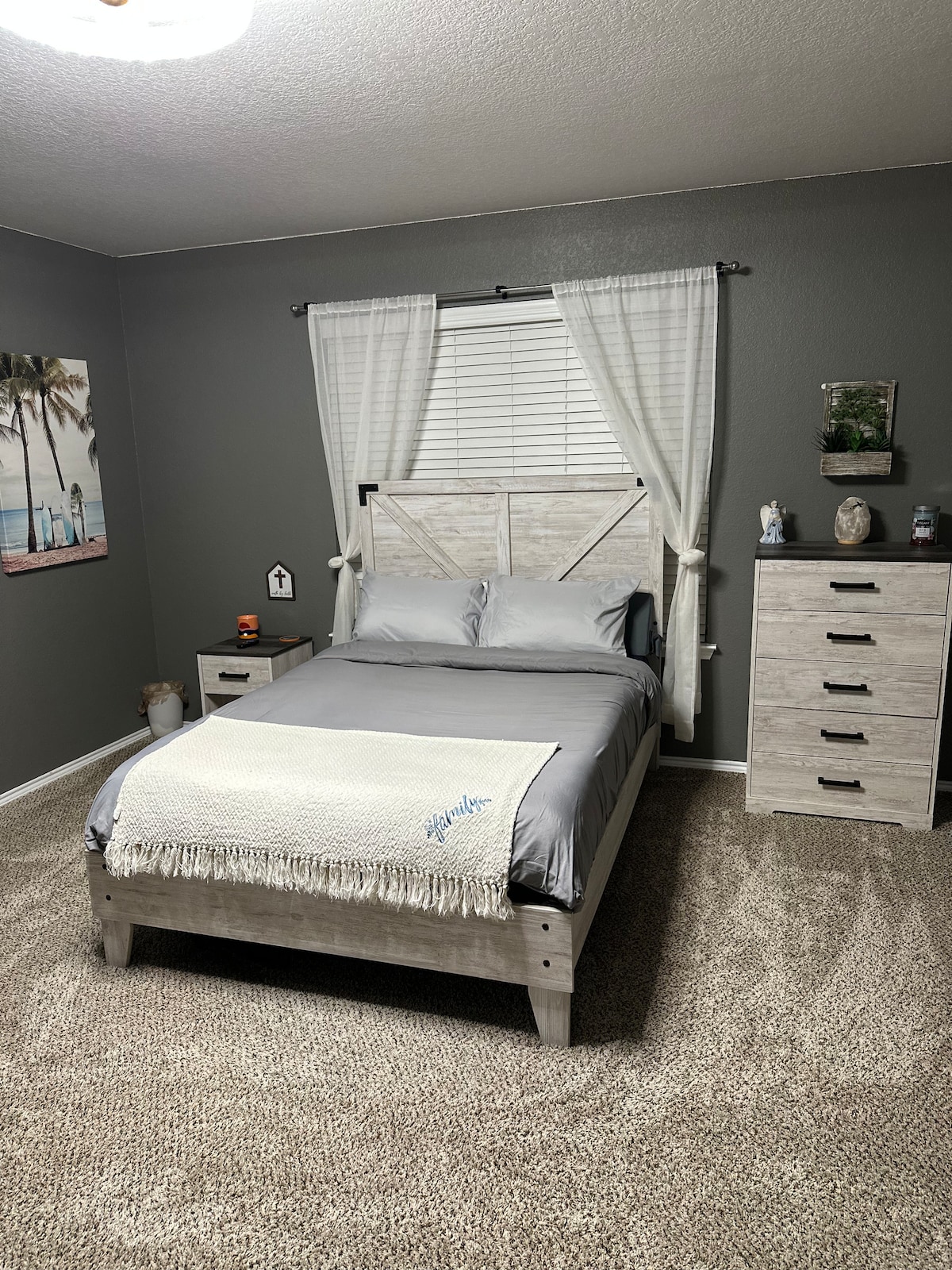 A welcoming bedroom features a light-colored wooden bed frame dressed in gray bedding. A cozy throw is draped at the foot of the bed. Natural light filters through the bright curtains, illuminating a textured carpet and a bedside table with a lamp. A dresser stands against the wall.