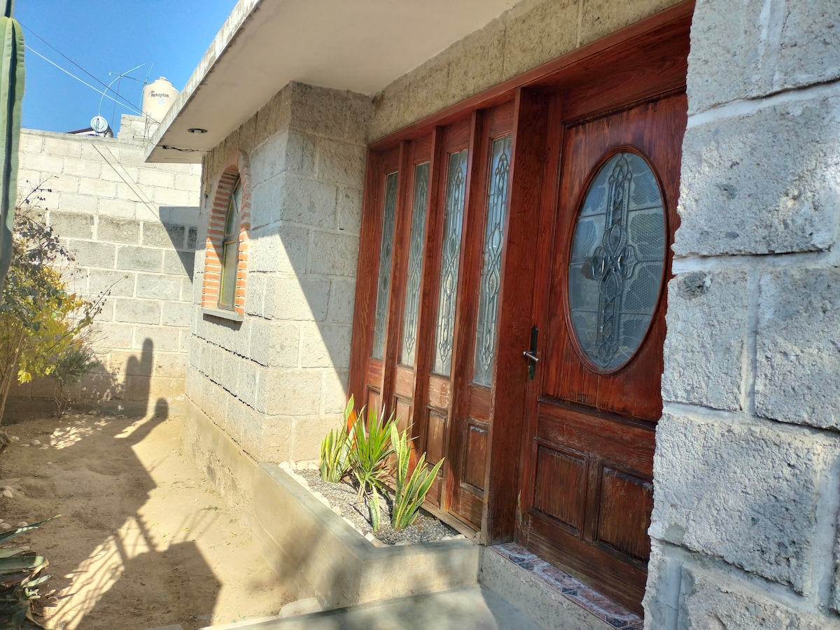 The entrance features large, decorative wooden doors with intricate glass insets. A small garden area with plants is situated beside the entrance, surrounded by a stone wall. The sunlight casts gentle shadows on the pathway, contributing to a serene outdoor setting.