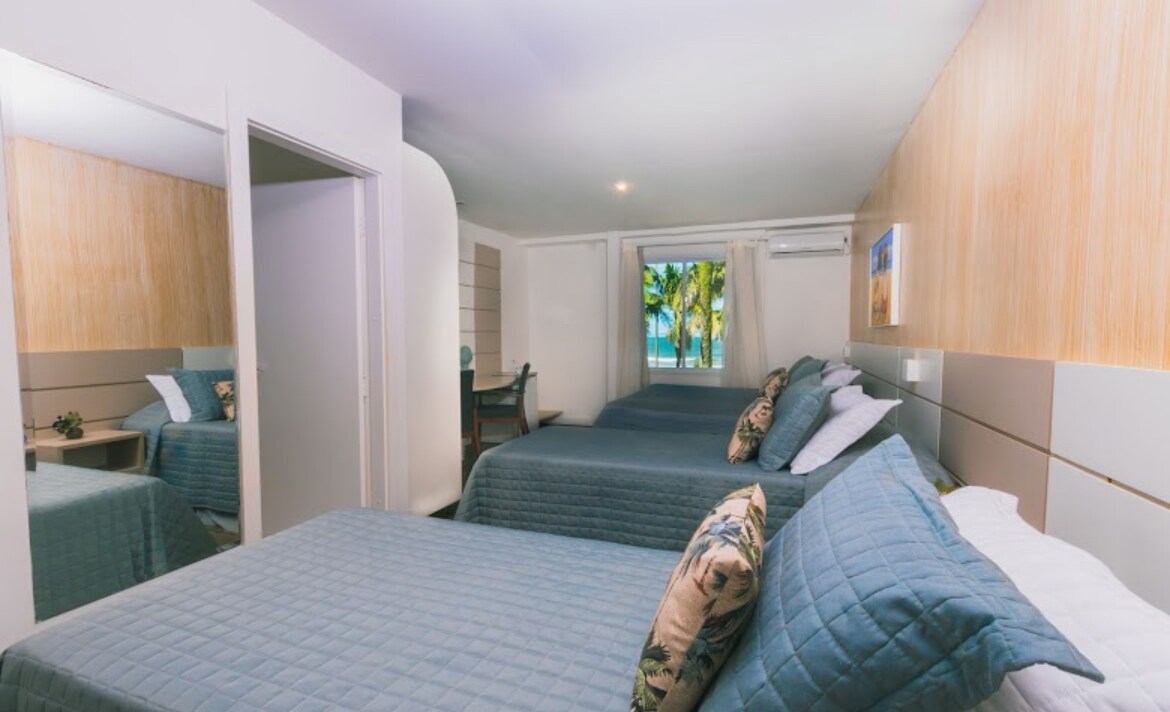 A well-organized room features two spacious beds, adorned with soft blue quilts and decorative pillows. Natural light is provided by a large window, revealing views of palm trees outside. A small desk is positioned beside the bed, and light-colored wooden accents enhance the room's design.