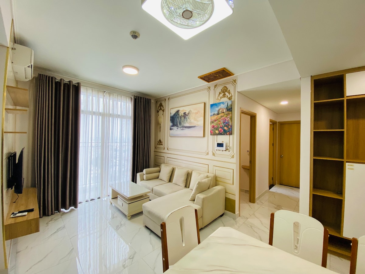 A bright living area features a light-colored sofa arranged around a coffee table. Floor-to-ceiling windows allow natural light to fill the space, complemented by soft curtains. Artwork decorates the walls, and a TV is positioned on a wooden stand, creating a comfortable gathering spot.