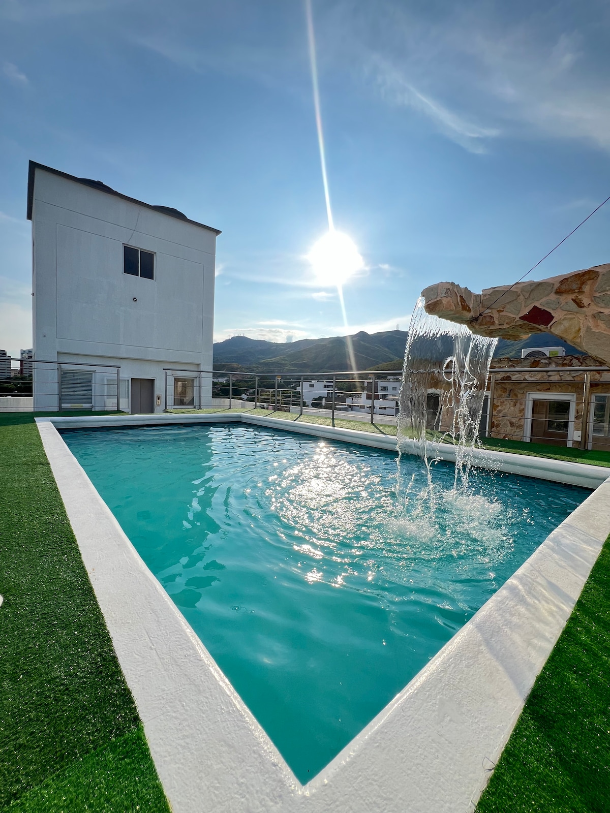 A rooftop pool glistens under the sun, with clear turquoise water and a gentle waterfall providing a soothing sound. The surrounding area features green artificial grass and a view of distant mountains, framed by a modern building structure in the background.
