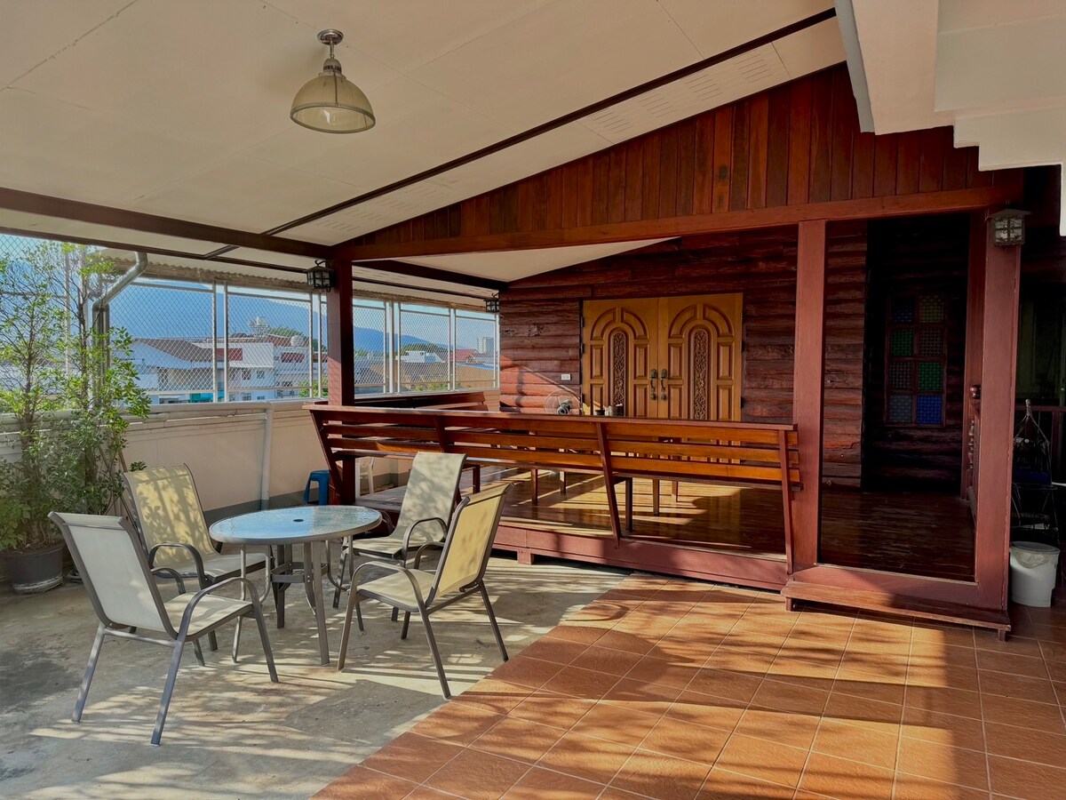 An outdoor seating area features a round glass table surrounded by four beige chairs. The wooden walls of the cabin extend the space, and large windows provide natural light. A decorative light fixture hangs from the ceiling, enhancing the open atmosphere.