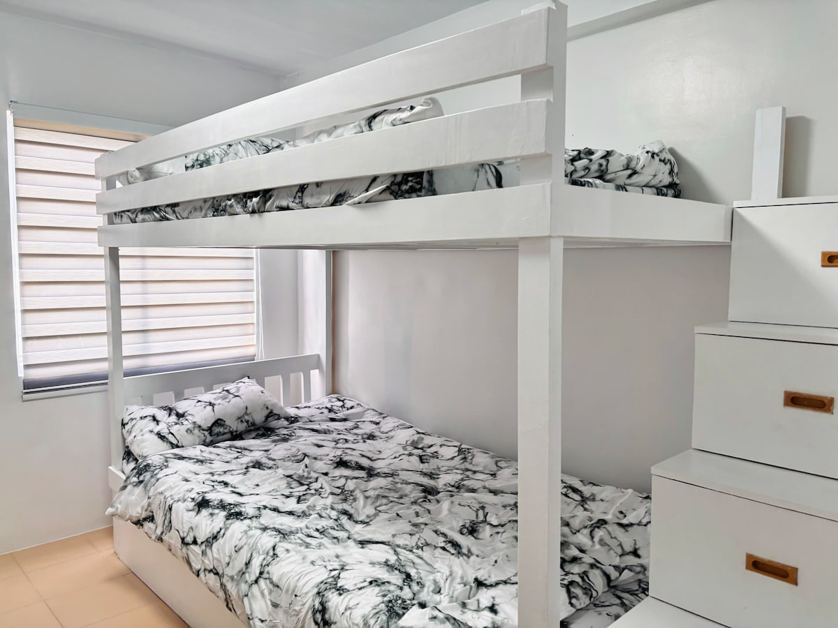 A tidy bedroom features a white bunk bed with a textured black-and-white bedding set. A nearby window offers natural light, and a matching dresser with drawers is positioned beside the bunk bed.