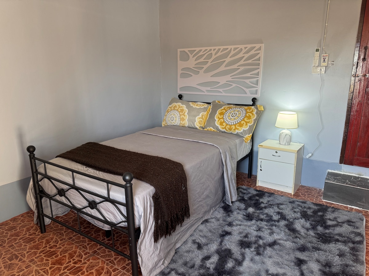 A bedroom features a metal bed frame with soft grey bedding adorned with decorative yellow pillows. A small bedside table holds a lamp providing gentle illumination. A cozy rug lies underfoot, and a simple headboard adds a modern touch to the space.