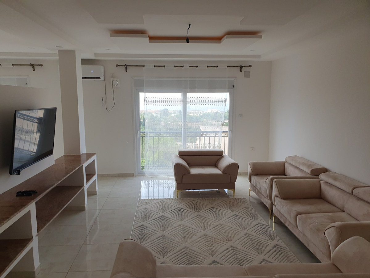 A modern living area features light-colored sofas arranged around a neutral rug. A sleek entertainment unit stands against the wall, while glass doors offer views of the outdoors, allowing natural light to fill the space. Air conditioning is also installed for comfort.