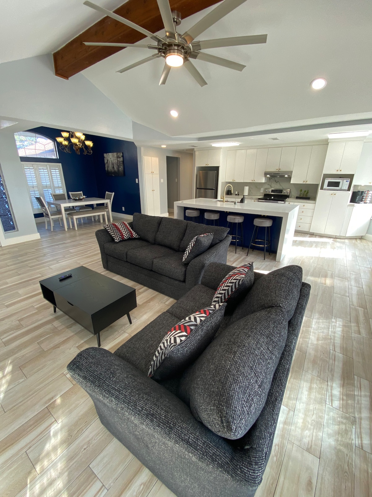 A spacious living area is presented with two dark gray couches adorned with patterned cushions. A sleek coffee table sits in the center. The kitchen area is visible in the background, featuring modern appliances and a dining table with six chairs, under a bright ceiling.