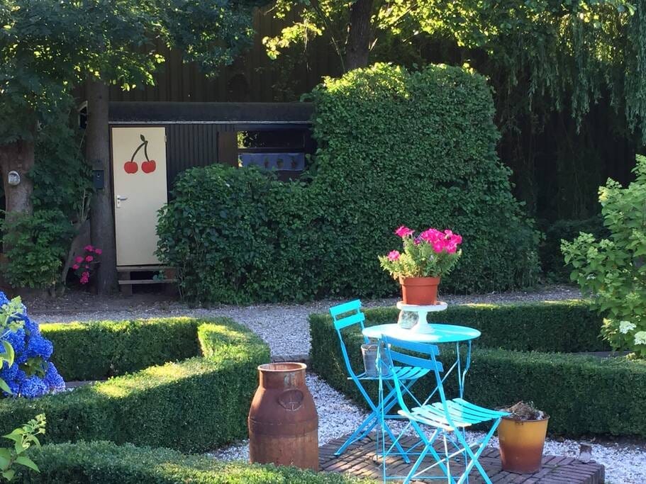 A charming outdoor area is presented, featuring neatly trimmed hedges and vibrant flowers. A small blue table with two matching chairs is positioned in front of a decorative pot filled with blooming flowers. A rustic metal container is included for added character.