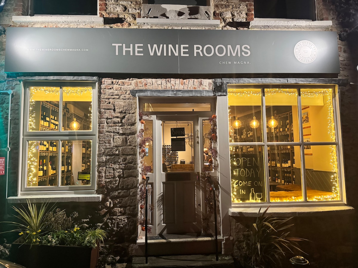 A charming entrance to The Wine Rooms is featured, illuminated by warm lights and festive decorations. The windows display various wine bottles, while the door is framed by glowing fairy lights, inviting guests to enter and explore the offerings inside.