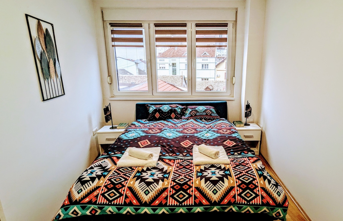 غرفة نوم مريحة مع سحر ريفي ومطبوعات حية على الفراش. يضيء المسكن بالضوء الطبيعي القادم من خلال نافذة كبيرة، مما يوفر جوًا دافئًا ومرحبًا للاسترخاء.