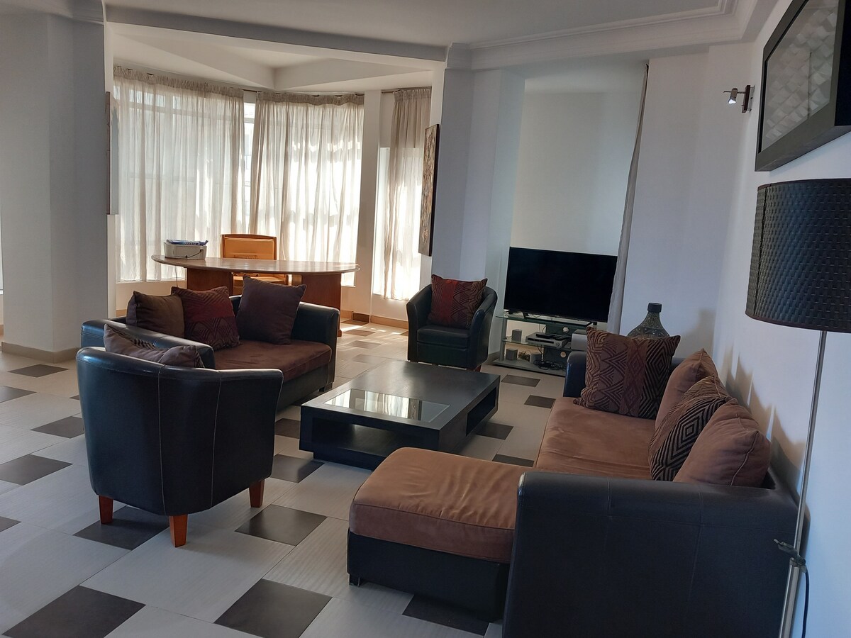 A spacious living area is furnished with brown sofas arranged around a low coffee table. An elegant dining table is visible in the background, accompanied by large windows that allow natural light to fill the room. A television and decorative accents complete the setting.
