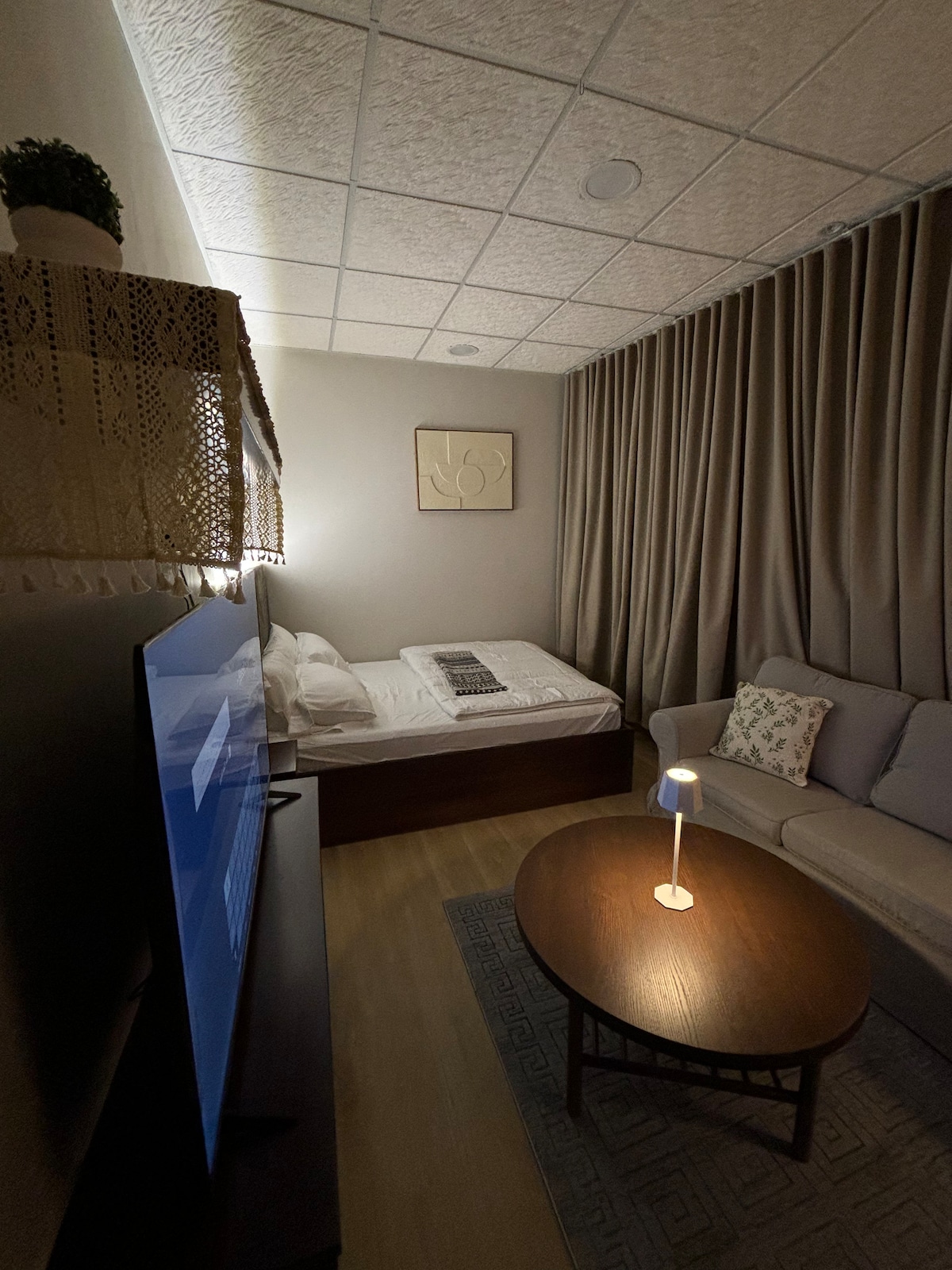 A cozy sleeping area is visible, featuring a bed positioned against a wall with soft bedding and a decorative throw. A comfortable sofa is placed near a round coffee table, illuminated by a small lamp. Elegant drapes frame the window area, contributing to the ambiance.