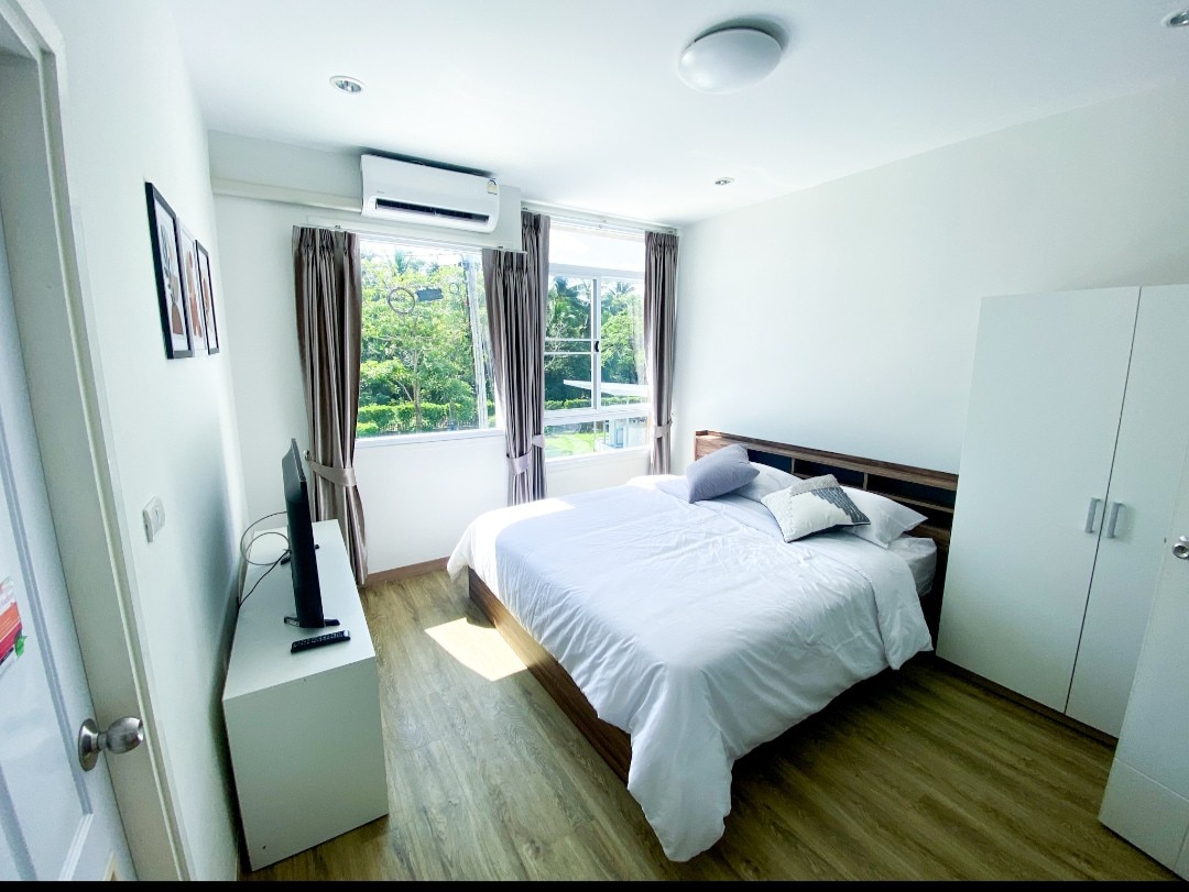 A well-lit bedroom features a spacious bed adorned with neutral bedding and decorative pillows. Large windows provide natural light and views of greenery outside. A flat-screen TV is positioned on a compact cabinet, and a wardrobe stands against the wall, contributing to the room's clean aesthetic.