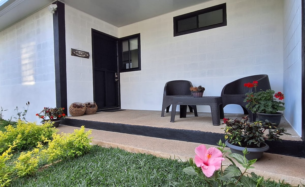 A welcoming front porch features two dark wicker chairs and a small table, surrounded by vibrant flower pots. The entrance is highlighted by a black door and window, while a neatly trimmed grassy area and blooming plants complete the serene outdoor space.