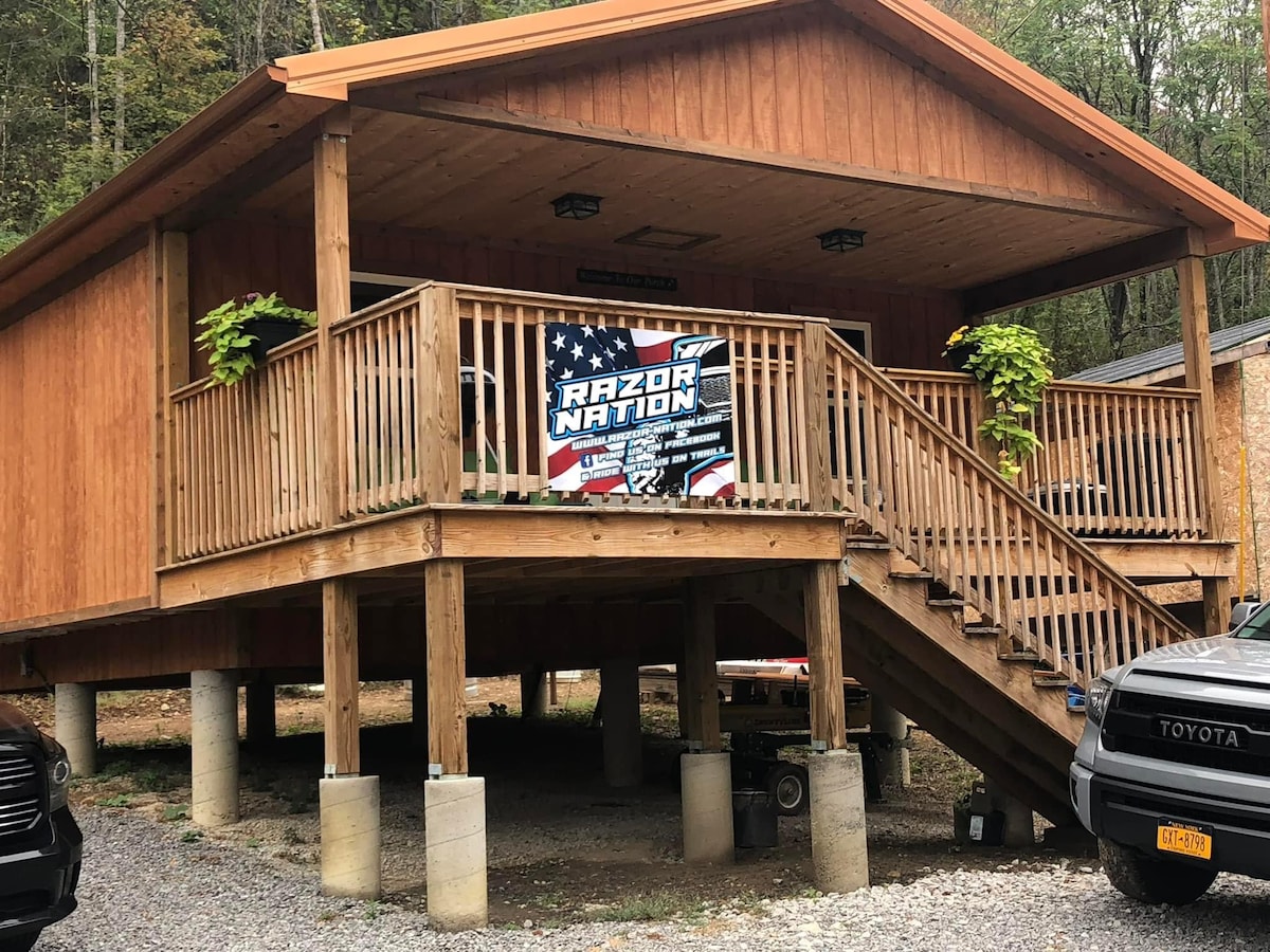 The spacious wooden cabin features a raised deck supported by concrete pillars. A large banner displays the words 'Razor Nation,' and potted plants are placed on the deck railing. A gravel area surrounds the cabin, which is bordered by trees.