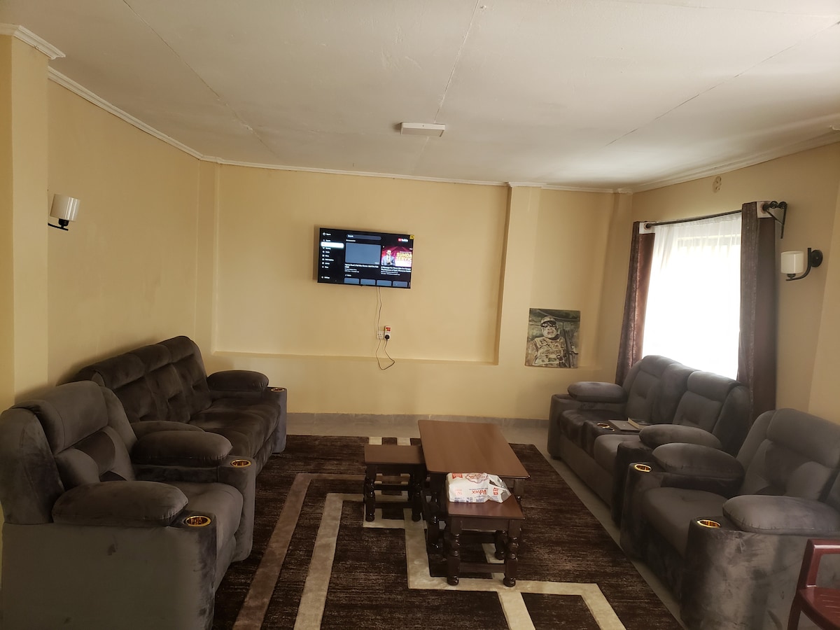 A cozy living room is furnished with plush gray sofas positioned around a central coffee table. A wall-mounted television is visible, providing entertainment options. Natural light enters through a window, complemented by dark curtains. A patterned area rug enhances the room's comfort.