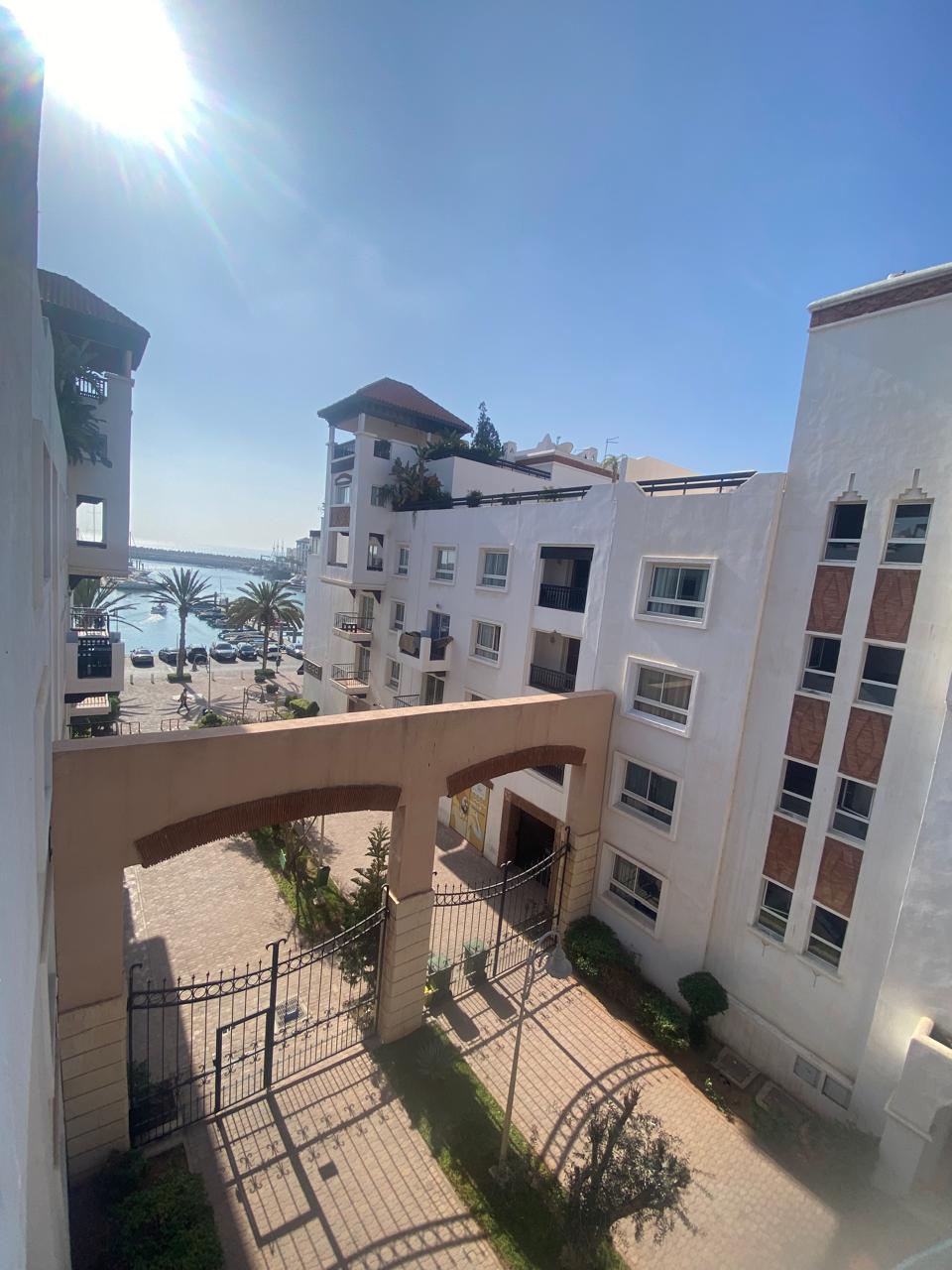 A view from a balcony showcases a well-maintained courtyard bordered by white buildings. Palm trees line the pathway, and a glimpse of the sea is visible in the background, enhancing the outdoor ambiance of the property.