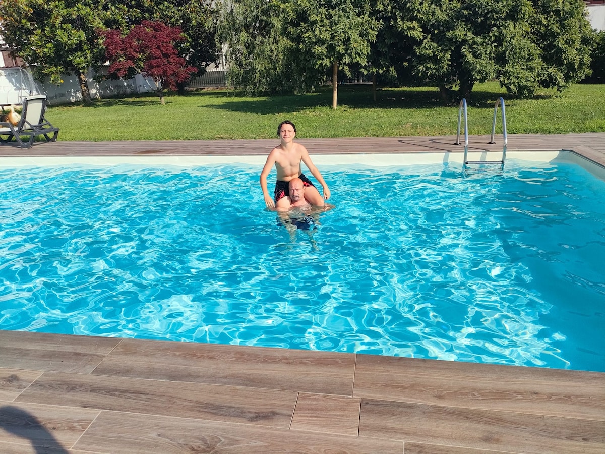 A crystal-clear pool is surrounded by a spacious wooden deck, reflecting the sun. Lush greenery is visible in the background, providing a serene setting for relaxation and recreation.