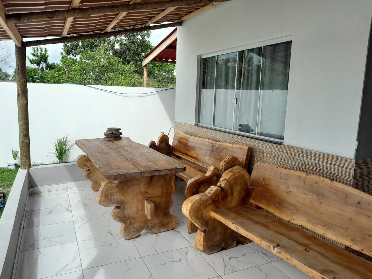An outdoor seating area features a large wooden table accompanied by rustic benches crafted from solid wood. Natural light filters in through the nearby window, and greenery is visible in the background, creating a relaxed setting.