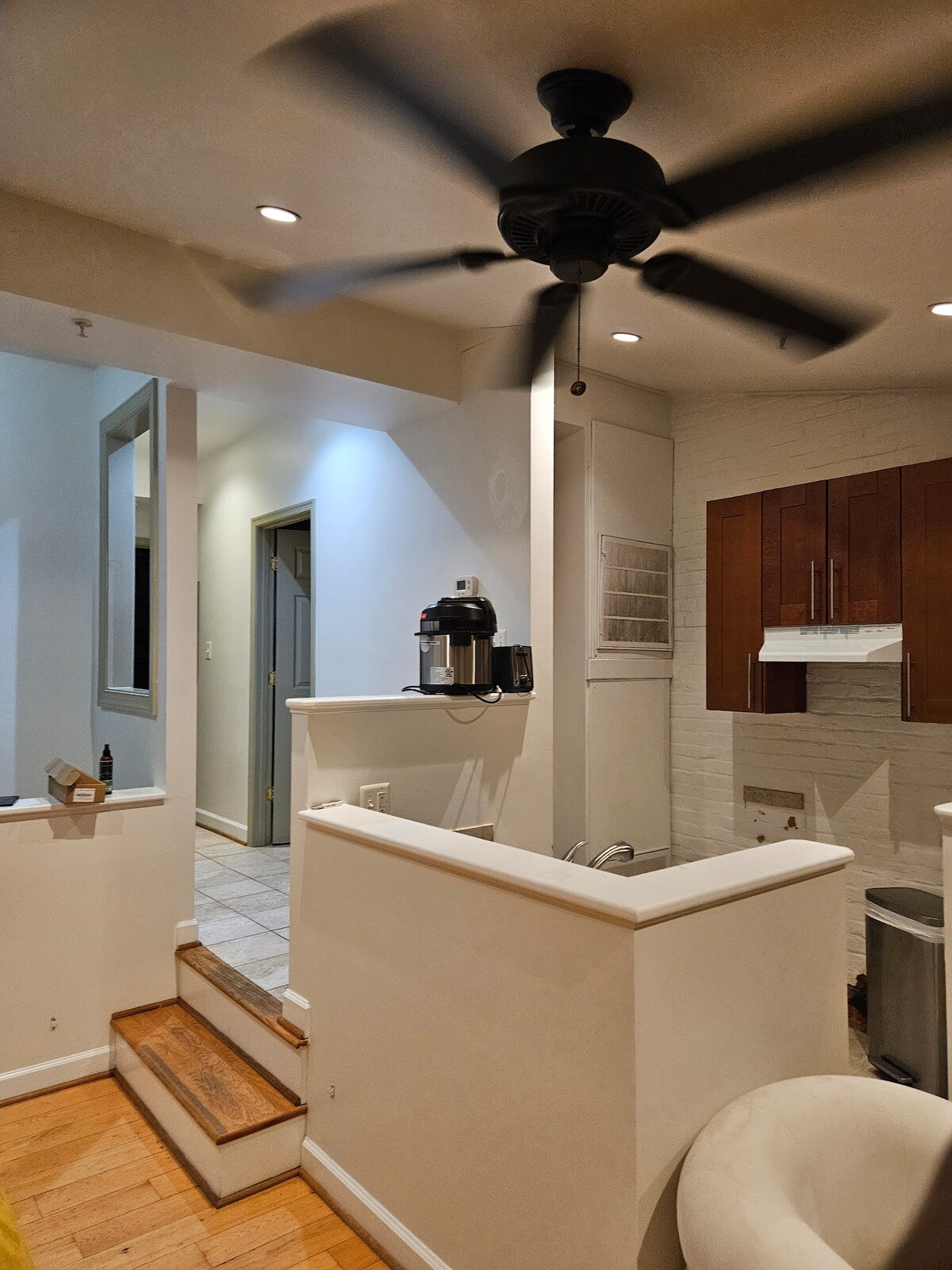 An open layout is seen, featuring a contemporary kitchen with wooden cabinetry. A ceiling fan provides ventilation above a central area, while a set of stairs leads towards a wider space. Soft lighting creates a welcoming ambiance throughout the room.