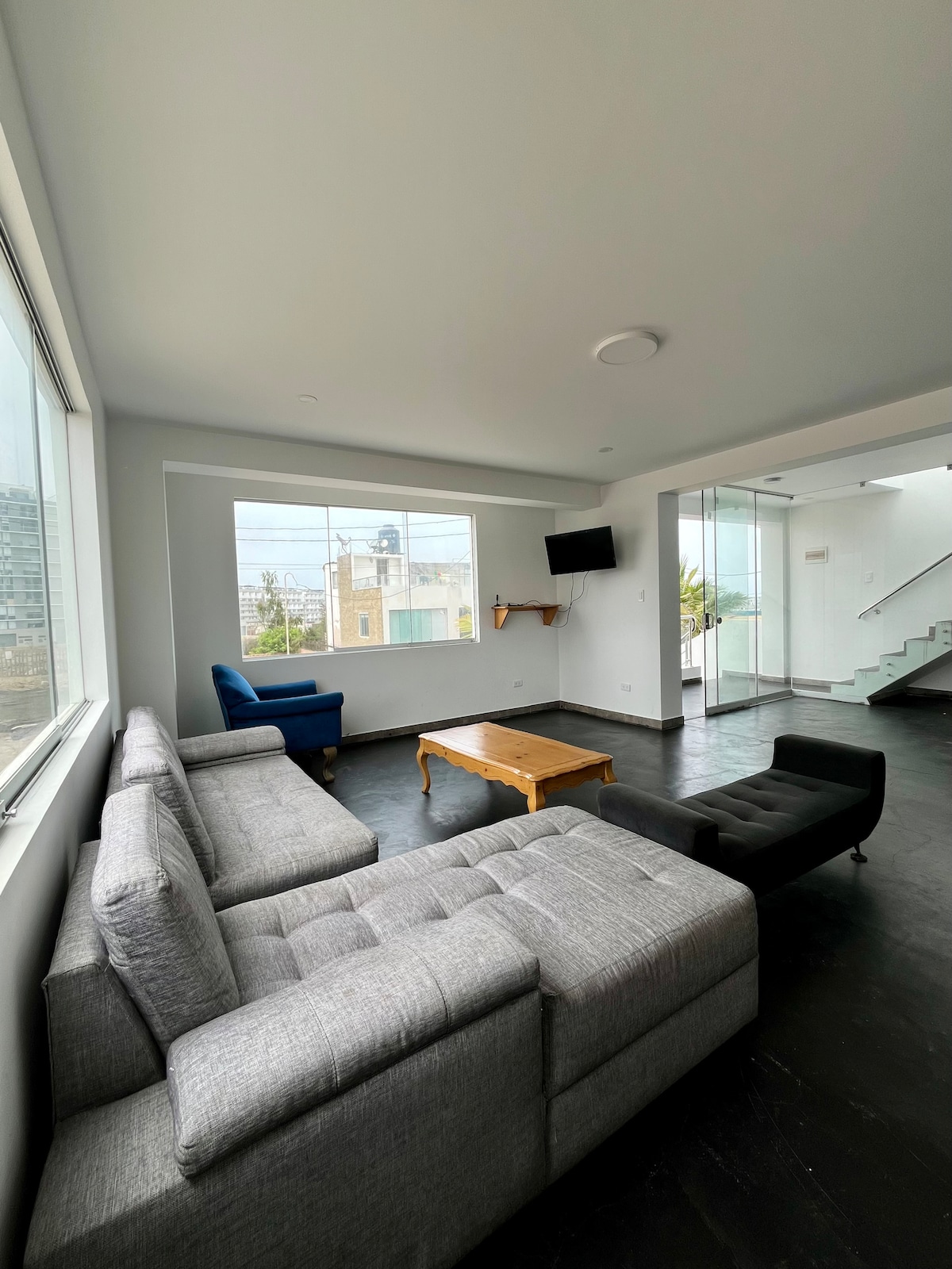 A bright living area is shown with a grey sectional sofa and a dark chair positioned near large windows. A wooden coffee table is centered in the room, with a wall-mounted TV visible. The space features neutral-colored walls and dark flooring, enhancing the modern design.