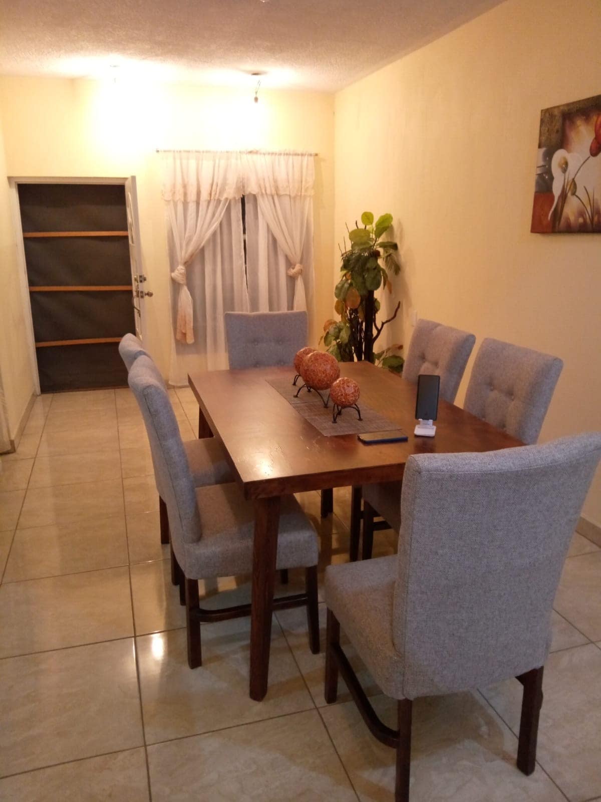 A dining area features a wooden table surrounded by six upholstered chairs. Natural light filters through sheer curtains at the window. A decorative plant is positioned in the corner, and artwork adorns the wall, contributing to a serene and inviting atmosphere.