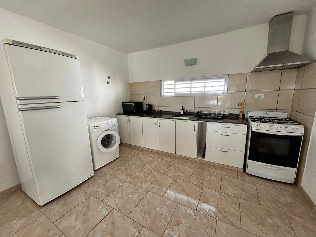 A functional kitchen features modern appliances including a refrigerator, stove, and washing machine. Light-colored cabinetry complements the tiled floor, while natural light floods in through a window, enhancing the spacious feel of the room.
