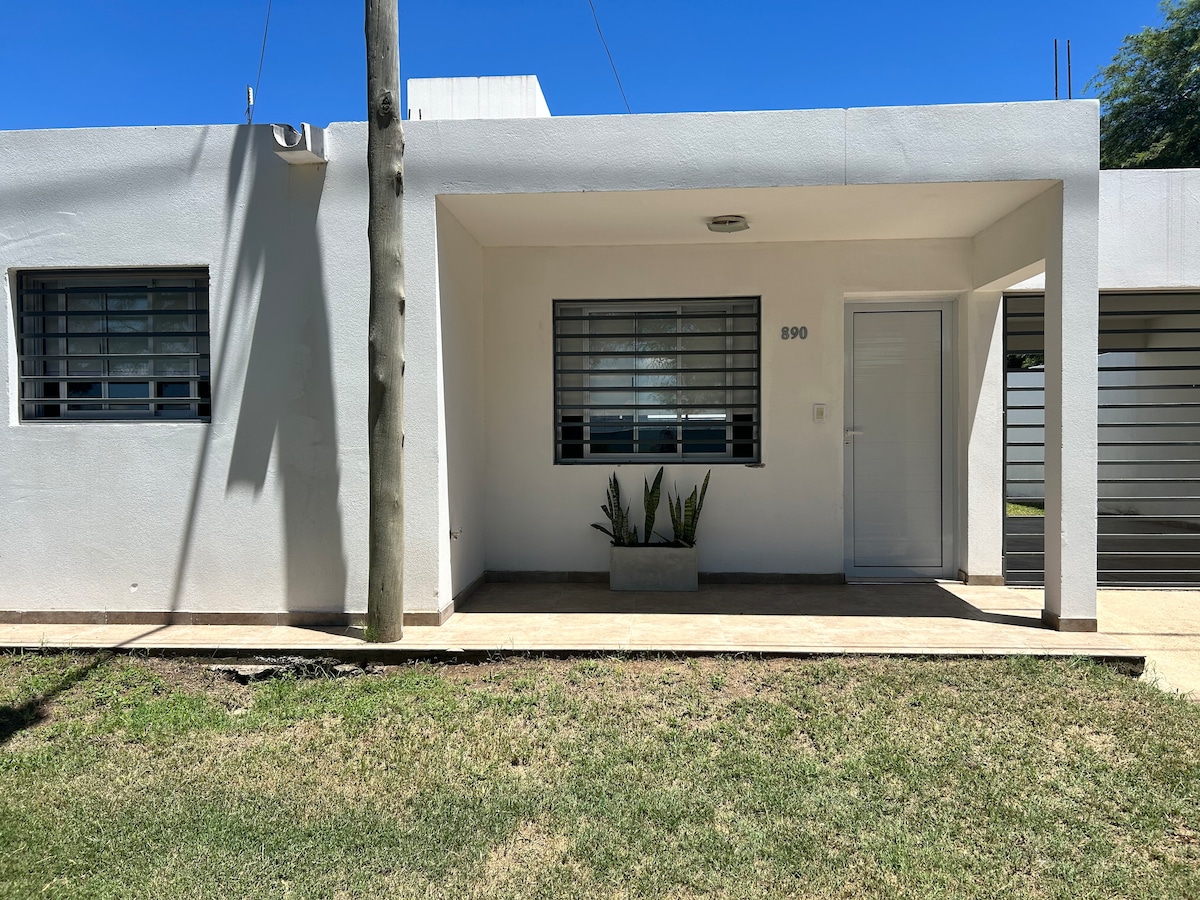 A modern white facade is featured, with large windows accented by metal grilles. A planter with greenery is placed near the entrance. The well-maintained lawn adds to the serene atmosphere, while the clear blue sky provides a bright backdrop.