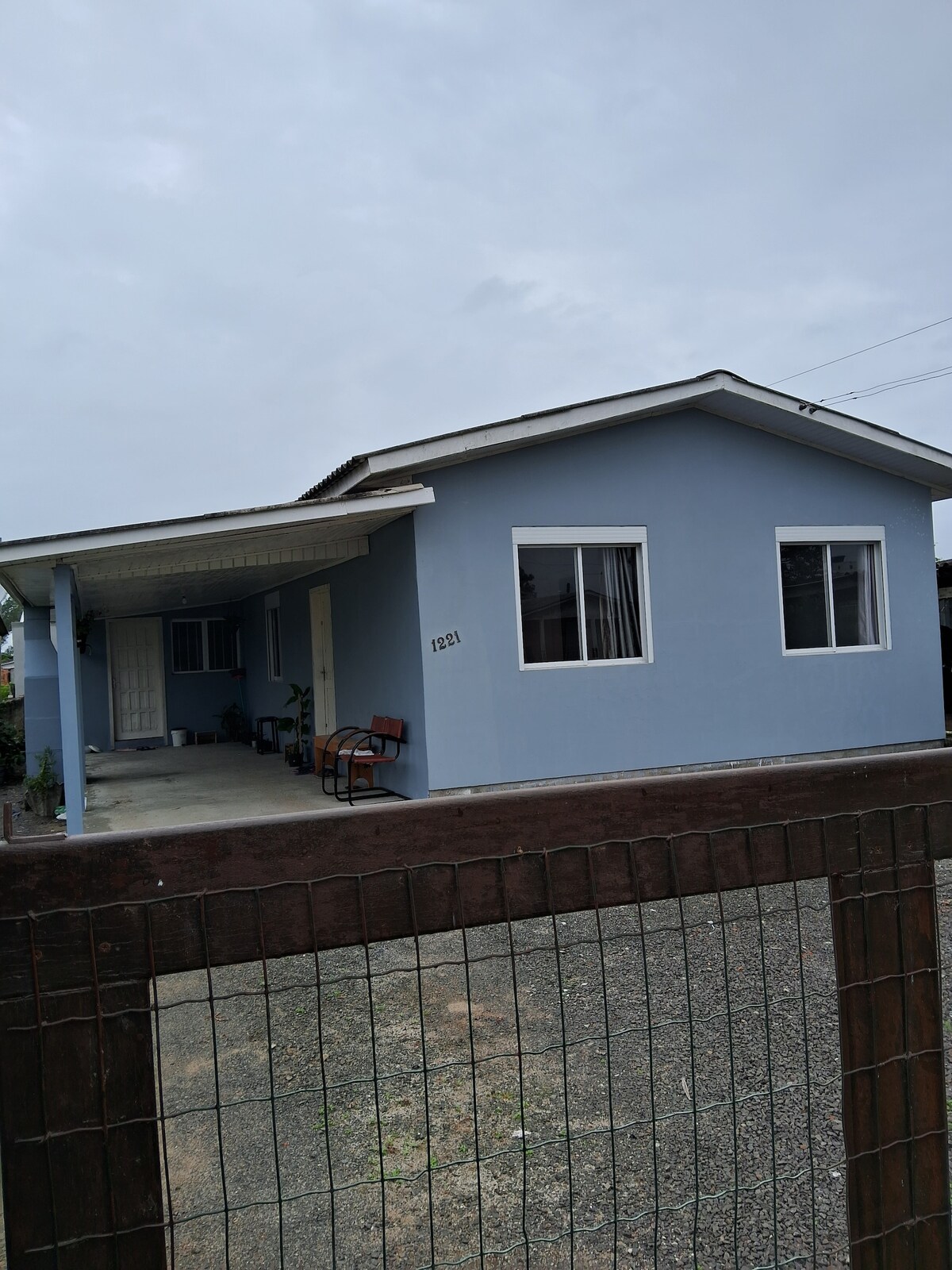A single-story blue house is depicted, featuring a covered porch with two wooden chairs positioned at the entrance. Large windows provide natural light to the interior. The property is enclosed by a simple fence, and a gravel driveway leads to the front of the house.