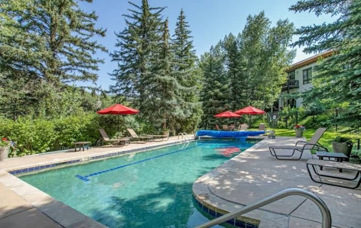 A spacious outdoor pool is surrounded by lush greenery and tall pine trees. Red umbrellas provide shade over lounge chairs situated along the pool's edge. The area offers a relaxing atmosphere for enjoying the outdoors and is accessible from the condo.