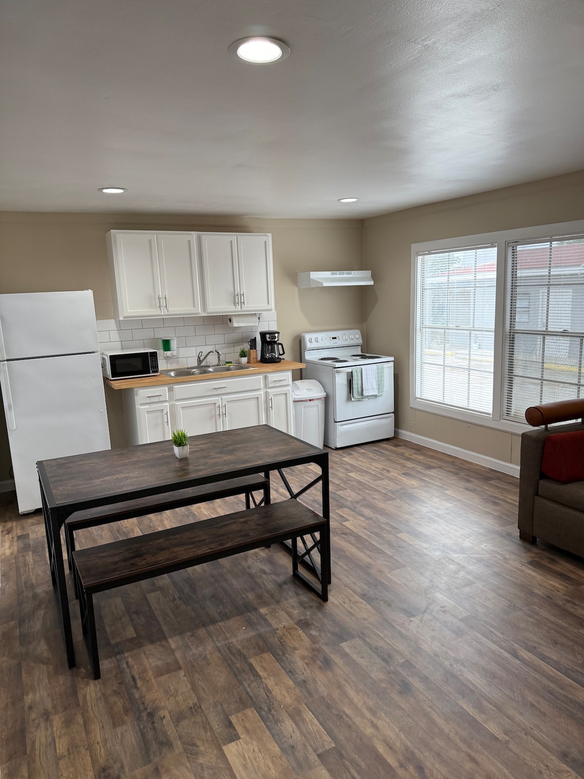 The kitchen area features modern appliances, including a refrigerator, microwave, and stove. A dark wood dining table with benches is positioned nearby, while natural light enters from two large windows, illuminating the space. Neutral-colored walls provide a simple backdrop.