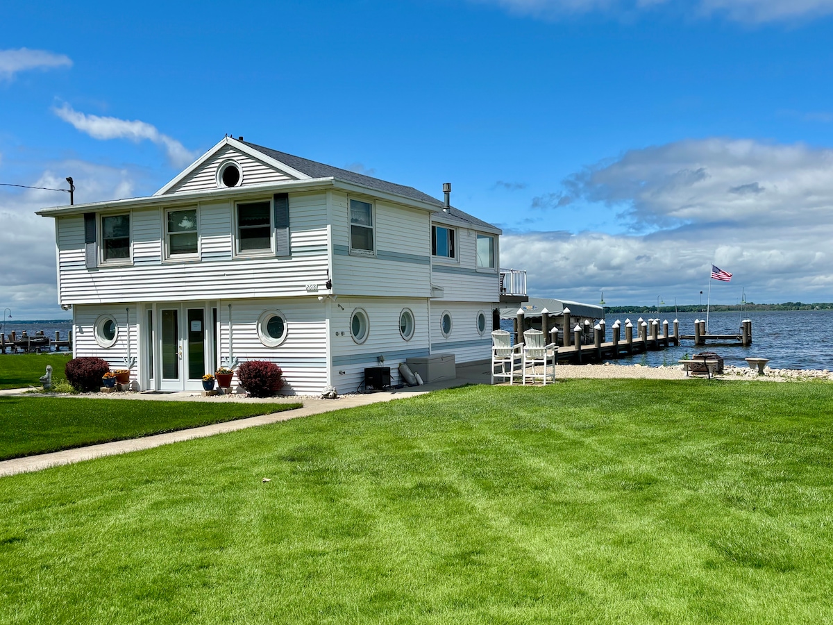 A two-story boathouse is depicted beside Lake Butte des Morts. Circular windows and a deck overlook the water, while a grassy area is maintained in front. A dock extends into the lake, with a flag visible in the background under a partly cloudy sky.